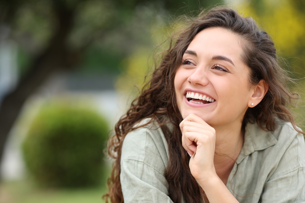 A joyful young woman smiling brightly in a natural outdoor setting, reflecting the benefits of restored teeth - Dental Implants Rancho Bernardo