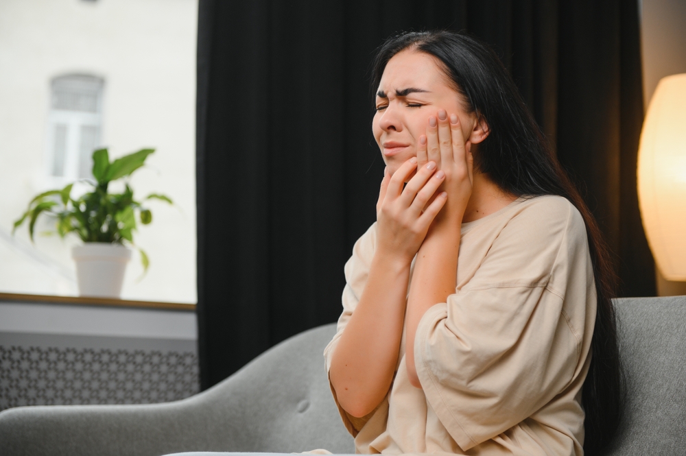 Woman in severe tooth pain needing emergency dentist - Rancho Bernardo Dentist Young woman holding jaw in pain while sitting on couch, showing need for urgent emergency dentist care - Rancho Bernardo Dentist