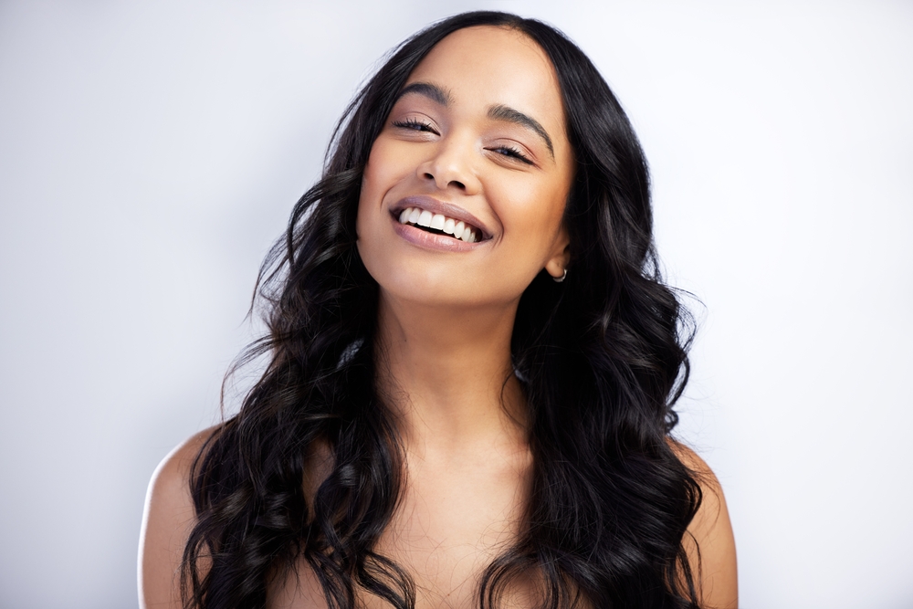 Smiling woman with bright teeth after dental crowns - Rancho Bernardo Dentist Confident young woman with long hair smiling against light background, representing dental crowns treatment - Rancho Bernardo Dentist
