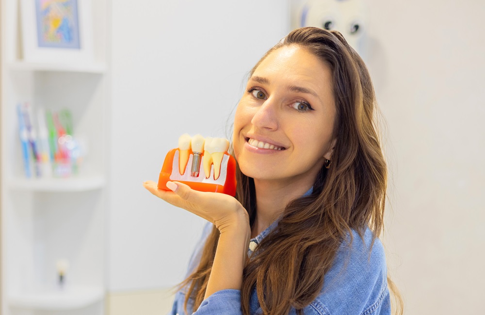 Smiling woman holding dental implant model - Anchor Dental & Implant Center Female patient showing dental implant model while smiling at dentist office - Anchor Dental & Implant Center