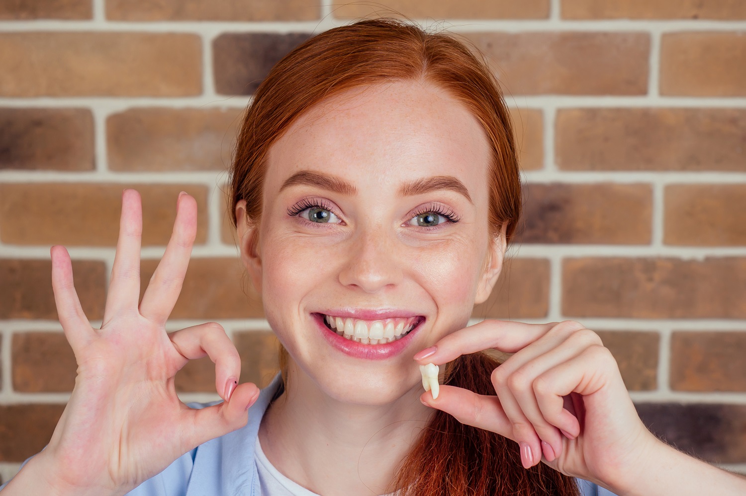 Smiling woman holding extracted tooth - Anchor Dental & Implant Center Happy young woman showing removed tooth and making okay gesture after tooth extraction - Anchor Dental & Implant Center