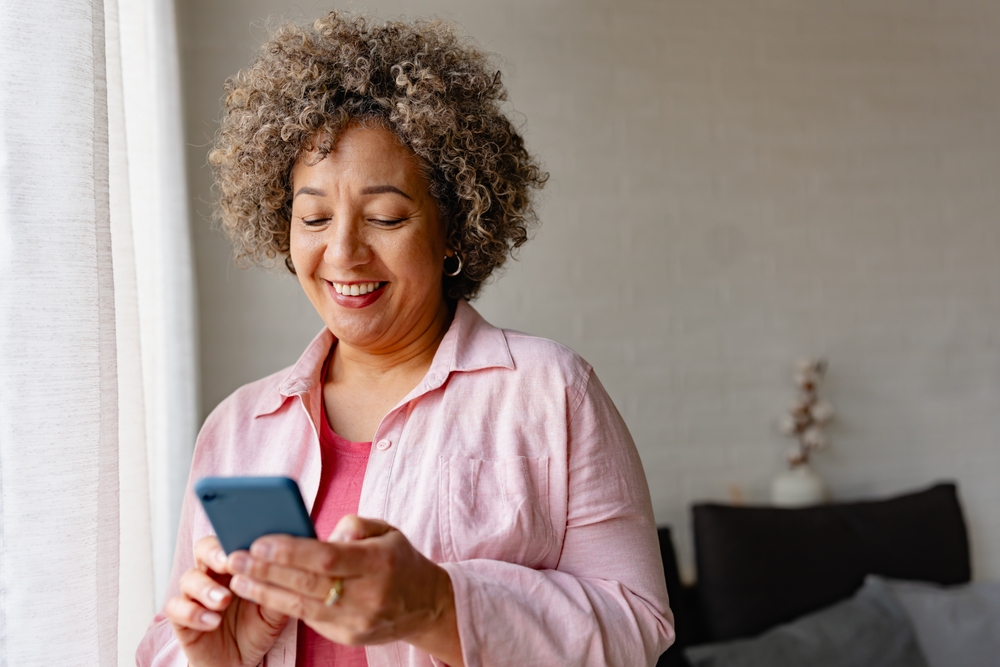 Smiling woman using phone at home after dental implants - Rancho Bernardo Dentist Middle-aged woman smiling while holding smartphone, representing restored confidence with dental implants - Rancho Bernardo Dentist