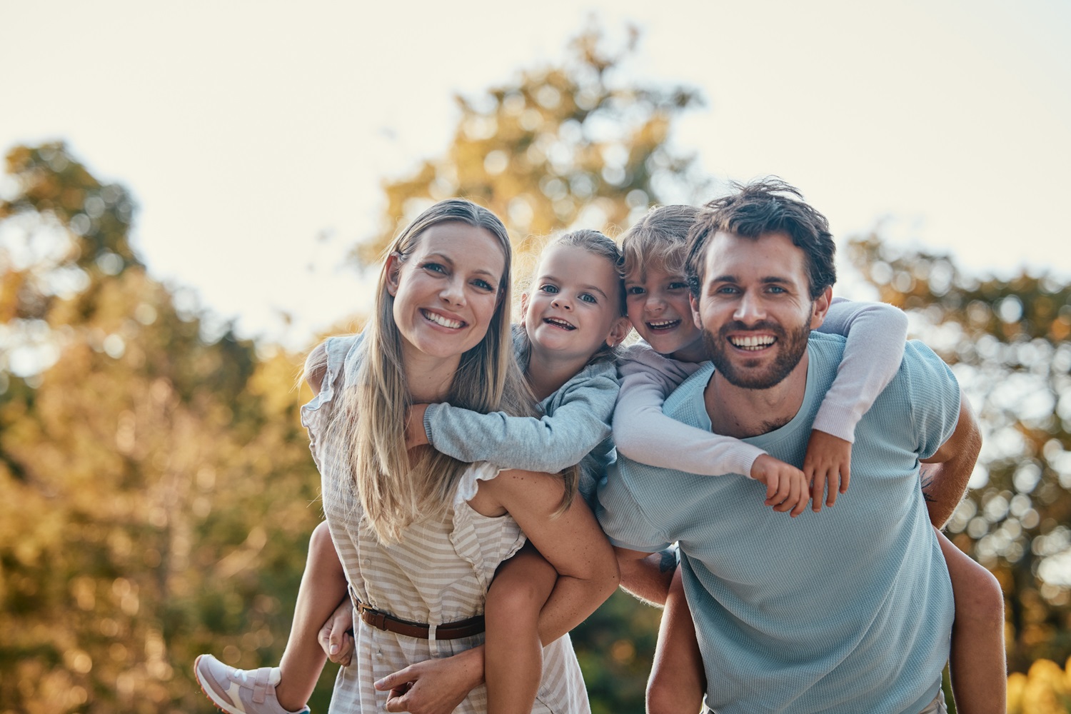 Smiling family enjoying quality time together outdoors – Anchor Dental & Implant Center Parents giving their two children piggyback rides while smiling happily in a park, symbolizing healthy family smiles – Anchor Dental & Implant Center