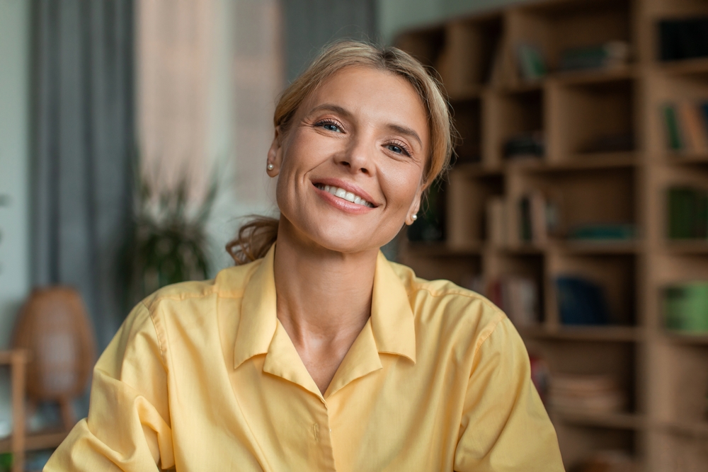 A woman in a yellow shirt smiling warmly indoors, highlighting her enhanced smile after teeth whitening - Teeth Whitening Rancho Bernardo