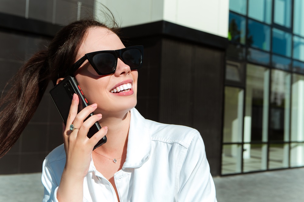 Smiling woman with perfect teeth after veneers treatment - Anchor Dental & Implant Center Young woman in sunglasses smiling while talking on phone, showing bright teeth from veneers - Anchor Dental & Implant Center